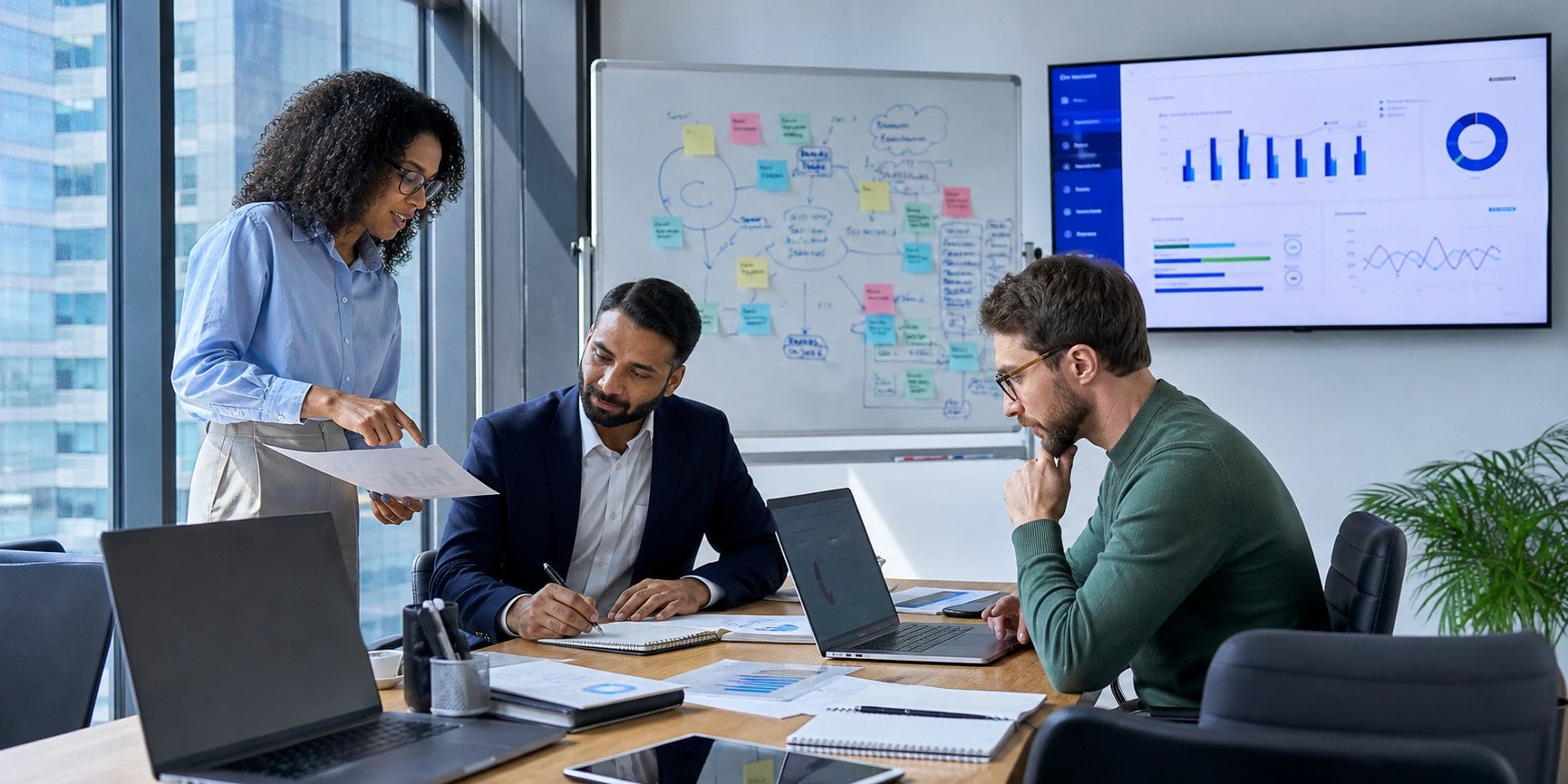 Three colleagues in a modern office reviewing an AI project together, with a whiteboard of colorful diagrams and a wall screen of data dashboards behind them
